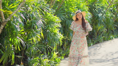 Cámara-Lenta-De-Una-Dama-Asiática-Caminando-Hacia-La-Cámara-Con-Un-Vestido-Largo-Y-Un-Sombrero-De-Paja-Sobre-Un-Fondo-Tropical-En-Bali
