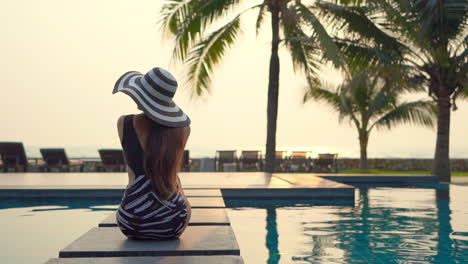 Back-view-of-a-slim-tourist-lady-sitting-near-a-pool,-wearing-a-black-and-white-swiming-suit-and-a-floppy-hat-,-admiring-the-view-during-sunset