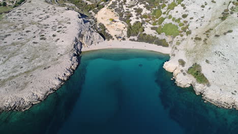Luftaufnahme-Einer-Kargen-Mittelmeerküste-Mit-Einem-Strand-Und-Dem-Kristallklaren-Meerwasser,-Bei-Dem-Man-Tief-Durch-Das-Wasser-Blicken-Kann