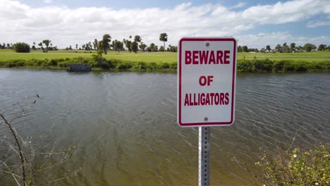 Beware-of-Alligators-Sign-by-Rippling-Pond-and-Green-Landscape,-Circling-Pan