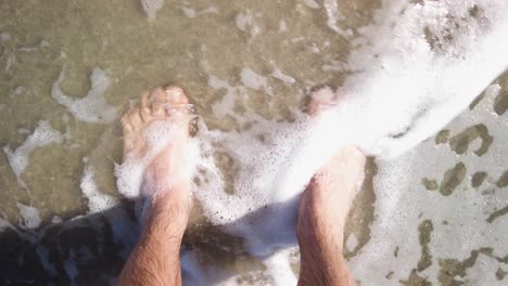 Foamy-crystalline-ocean-wave-washes-over-male-feet,-Slowmo-Top-Down-Shot