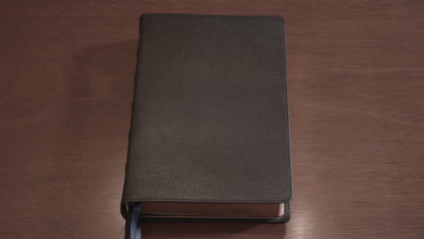 Hand-öffnet-Ein-In-Leder-Gebundenes-Heiliges-Bibelbuch,-Das-Auf-Einem-Holztisch-Sitzt