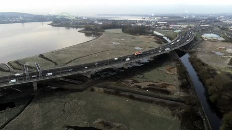 River-marsh-land-coast-aerial-push-in-over-commuter-traffic-crossing-bridge-uk