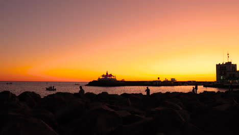Silhouette-fishermen-on-pier-and-coast-during-orange-sunset-casting-fishing-line-on-the-open-sea-in-the-background-is-the-city-and-harbor-of-sunset-Grand-Canary-island-4k-slow-motion-capture-at-60fps