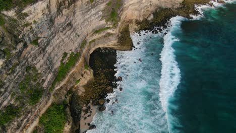 Aerial-view-ascending-shot,-waves-hitting-the-rocky-shore-of-the-Tanjung-Ringgit-cliff,-green-grass-and-trees-in-the-background