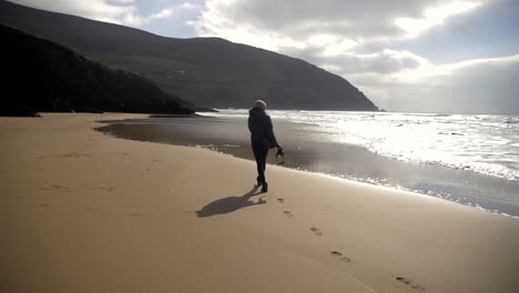 Female-photographer-walks-on-sandy-beach-in-Ireland,-leaving-footsteps-behind,-waves-hitting-the-shore,-sunny-sky-with-clouds,-slow-motion-shot