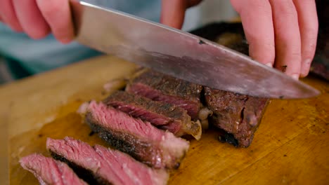 Close-up-view-of-Male-hand-with-a-knife-slicing-the-Grilled-Beef-Steak-on-cutting-Board-on-Wooden-Background