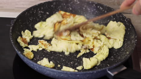 Dry-scrambled-eggs-in-black-skillet-stirred-with-wood-spatula,-close-up