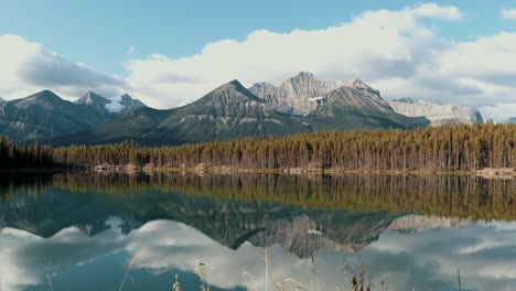 A-beautiful-timelapse-of-a-mountain-somewhere-in-Banff,-with-a-lake-and-the-reflection-of-the-mountains-showimng-on-the-calm-water-of-the-lake,-with-clouds-passing-by-and-the-sun-peaking-trough-them