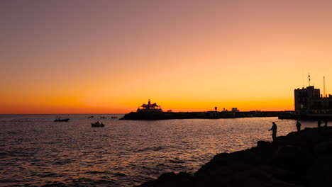 Fischersilhouette-An-Steiniger-Küste-Während-Des-Orangefarbenen-Sonnenuntergangs-Angelschnur-Auf-Offener-See-Im-Hintergrund-Ist-Stadt-Und-Hafen-Des-Sonnenuntergangs-Grand-Canary-Island-Valley-4k-Zeitlupenaufnahme-Mit-60fps