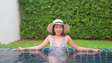 Standing-in-the-warm-water,-a-young-Asian-woman-leans-against-the-back-wall-of-a-resort-pool-smiling-at-all-she-can-see