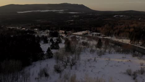 Aerial-drone-footage-of-a-beautiful-sunset-over-a-village-during-the-Canadian-winter-in-Quebec-with-golden-hour-colors-everywhere-and-the-sun-over-the-horizon,-as-well-as-beautiful-trees-and-snow
