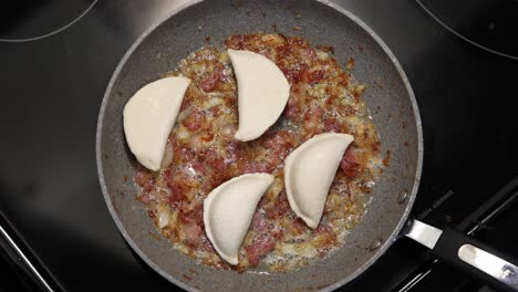 Gloved-hands-add-perogies-to-a-pan-of-sizzling-bacon-on-black-stovetop
