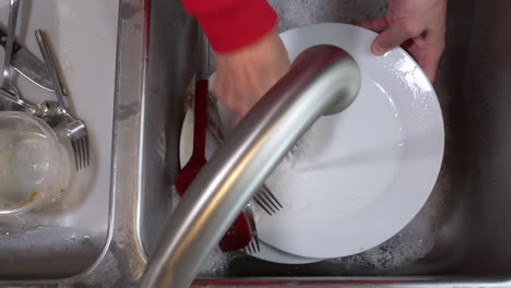 Top-down-view,-faucet-running,-cleaning-dirty-plates-and-spatula-in-sink-with-with-palm-dish-brush
