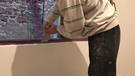 Young-man-removing-excess-of-plaster-with-scraper-around-wooden-window-frame-with-old-brick-wall-in-background-in-4k