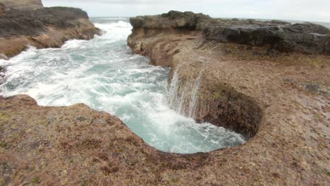 A-surge-of-ocean-water-as-it-pushes-up-into-a-hole-in-the-rock-shelf-and-crashes-back-down
