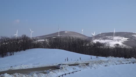 Viele-Windkraftanlagen-Sind-Auf-Dem-Schneebedeckten-Berg-Zu-Sehen,-Während-Im-Vordergrund-Zwei-Autos-Vorbeifahren,-Südkorea