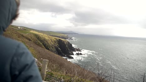 Person-in-green-hooded-jacket-stands-on-edge-and-watches-towards-cliffs-and-wild-sea-in-distance,-waves-crash-on-shore,-tourist-in-Dingle-peninsula,-Ireland,-on-cloudy,-windy-day,-slow-motion