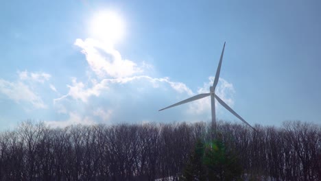 Closeup-view-of-the-wind-power-turbine-propeller-behind-trees-and-bright-sun-on-the-cloudy-sky-in-Pyeongchang,-South-Korea