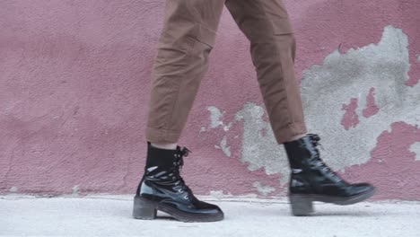 Slow-motion-close-up-of-a-woman-walking-forward-wearing-shiny-black-boots-and-brown-pants-at-an-urban-set-up-with-a-red-wall-as-a-background