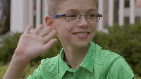 Cute-boy-with-glasses-sitting-outside-in-front-yard-looks-down-street,-smiles-and-waves-in-slow-motion