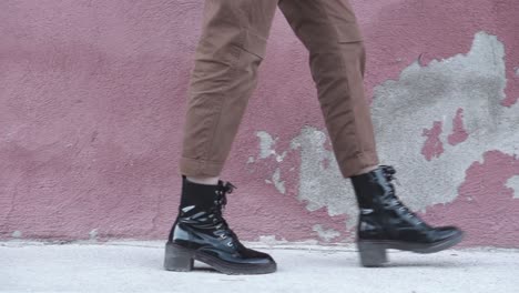 Close-up-of-a-woman-walking-forward-wearing-shiny-black-boots-and-brown-pants-at-an-urban-set-up-with-a-red-wall-as-a-background