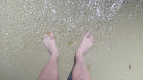 A-point-of-view-shot-looking-down-at-a-mans-feet-while-small-waves-lap-at-his-toes