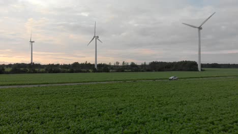 Arieal:-Autofahrt-Durch-Felder-Bei-Sonnenuntergang-Mit-Windrädern-Im-Hintergrund