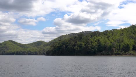 Slow-pan-across-calm-lake-with-rolling-forest-hills-on-sunny-day-with-cumulus-clouds