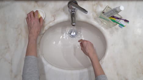 Badezimmer-Mit-Zahnbürsten---Die-Hand-Der-Alten-Frau-Wird-Auf-Leitungswasser-Mit-Seife-Gewaschen,-Dann-Gespült-Und-Auf-Einem-Weißen-Stoffhandtuch-Abgewischt---Draufsicht-Statische-Nahaufnahme-4k