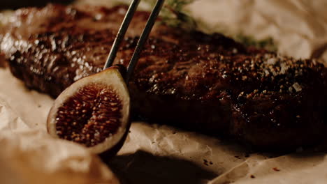 Placing-fig-with-kitchen-tongs-as-garnish-next-to-cooked-ribeye-steak,-close-up