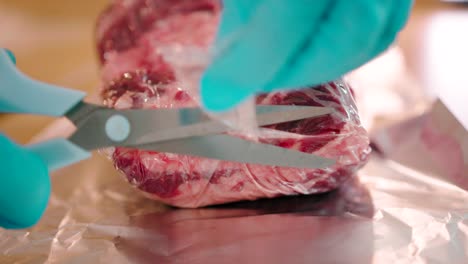 Close-up-view-of-a-man-cutting-the-steak-packed-polythene-wrap-with-a-pair-of-scissors