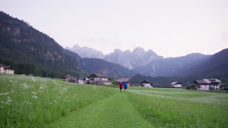 Two-hikers-walking-towards-a-village-with-mountains-in-the-background-at-sunset