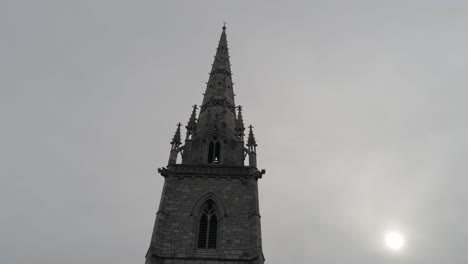 Aerial-view-rising-tilt-down-of-decorative-medieval-stone-church-tower-steeple-against-overcast-sky