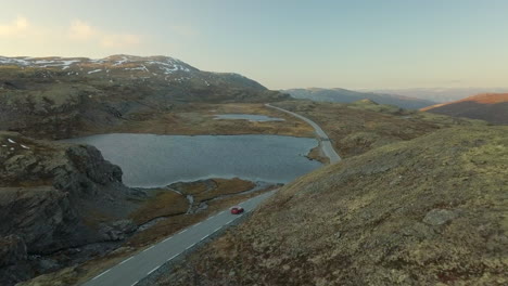 Coche-Rojo-Brillante-Conduciendo-Por-Una-Carretera-Estrecha-Y-Vacía-En-Una-Montaña-árida-En-Noruega,-Tiro-De-Drones-Con-Movimiento-Hacia-Adelante