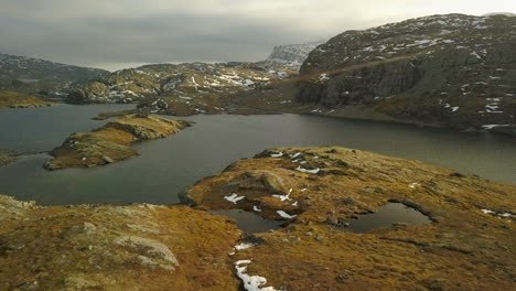 Hombre-Corriendo-En-La-Cima-De-Una-Montaña-En-Noruega-Con-Lagos-Y-Nieve,-Movimiento-De-Drones-Hacia-Adelante