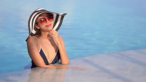 Attractive-asian-woman-wearing-bikini,-big-sunglasses-and-a-striped-black-and-white-floppy-hat,-standing-at-the-edge-of-a-swimming-pool