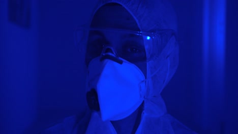 Man-wears-protective-suit,-face-mask-and-goggles,-looks-anxiously-around-dark-misty-room-with-flashing-red-blue-light