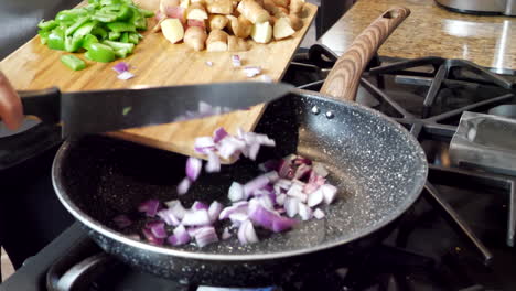 Pushing-red-onions-and-peppers-into-hot-pan-to-cook,-potatoes-on-wooden-cutting-board
