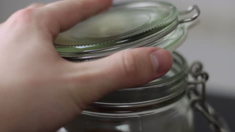 Close-up-of-opening-the-clasp-on-a-glass-mason-jar-and-lifting-the-lid-before-tilting-the-jar-forwards