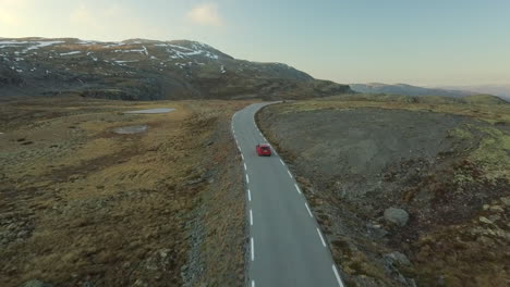 Coche-Rojo-Brillante-Conduciendo-Por-Una-Carretera-Estrecha-Y-Desierta-En-Una-Montaña-árida-En-Noruega,-Tiro-De-Drones-Con-Movimiento-Hacia-Adelante