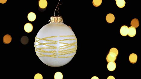Single-White-Christmas-Ball-Ornament-Mit-Goldenen-Glitzerstreifen-Und-Unscharfen-Lichtern,-Die-Im-Hintergrund-Flackern,-Nahaufnahme
