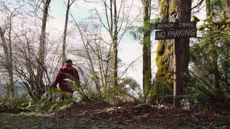 Man-walking-in-a-west-coast-rain-forest-on-Vancouver-Island-Canada-with-Beach-trail-sign-on-a-tree-leading-to-the-beach