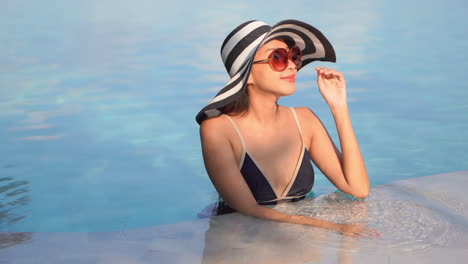 A-young-pretty-woman-stands-in-the-shallow-end-of-a-resort-pool-adjusts-her-sunglasses-as-she-looks-around
