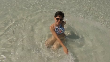 A-happy-girl-in-swimming-suit-playing-around-with-her-hands-in-the-sea-while-sitting-at-the-bottom-of-the-white-sand