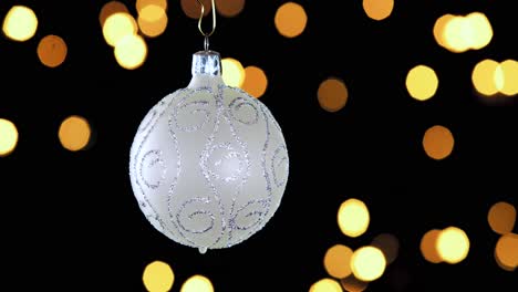 Single-White-Christmas-Ball-Ornament-Mit-Silbernen-Glitzerstreifen-Und-Unscharfen-Lichtern,-Die-Im-Hintergrund-Flackern,-Nahaufnahme