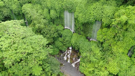 Aerial-orbital-shot-around-a-high-waterfall-in-a-lush,-green-jungle,-water-falling-forming-a-curtain-than-a-watercourse-on-rocks-continue-as-a-river