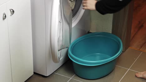 Barefoot-woman-placing-a-bucket-under-a-washing-machine-before-opening-it-and-unloading-clothes-into-it