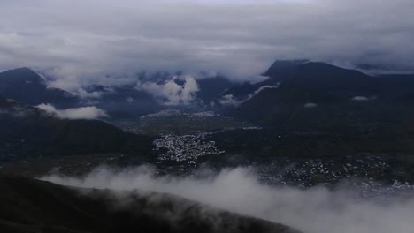 Dark,-gloomy-aerial-shot-as-flying-over-a-ridge-fog-whirling-around-as-clouds-covering-mountain-top-around-a-mall-city-in-the-valley