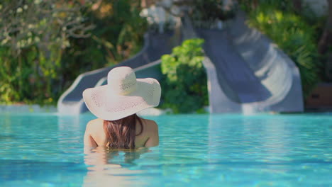 Woman-standing-in-swimming-pool-wearing-large-hat-facing-water-slides-surrounded-by-lush-tropical-landscaping